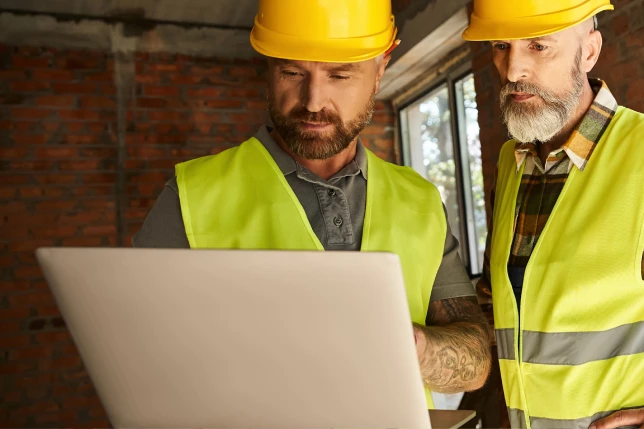 Two construction workers in shade looking at a laptop on a construction site